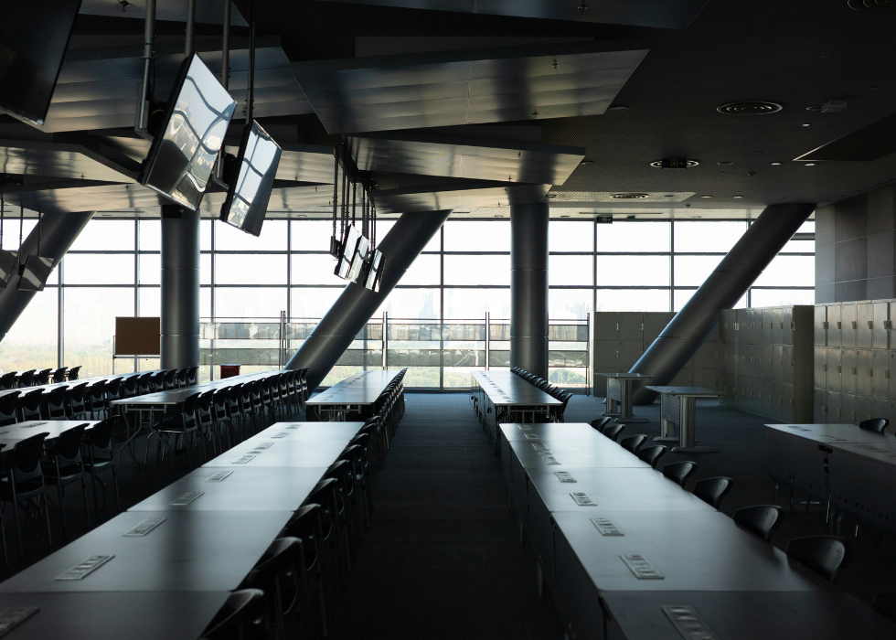 Modern classroom with long tables and chairs in rows, large windows along one side letting in daylight, clean minimalist interior, set up for group learning and presentations.
