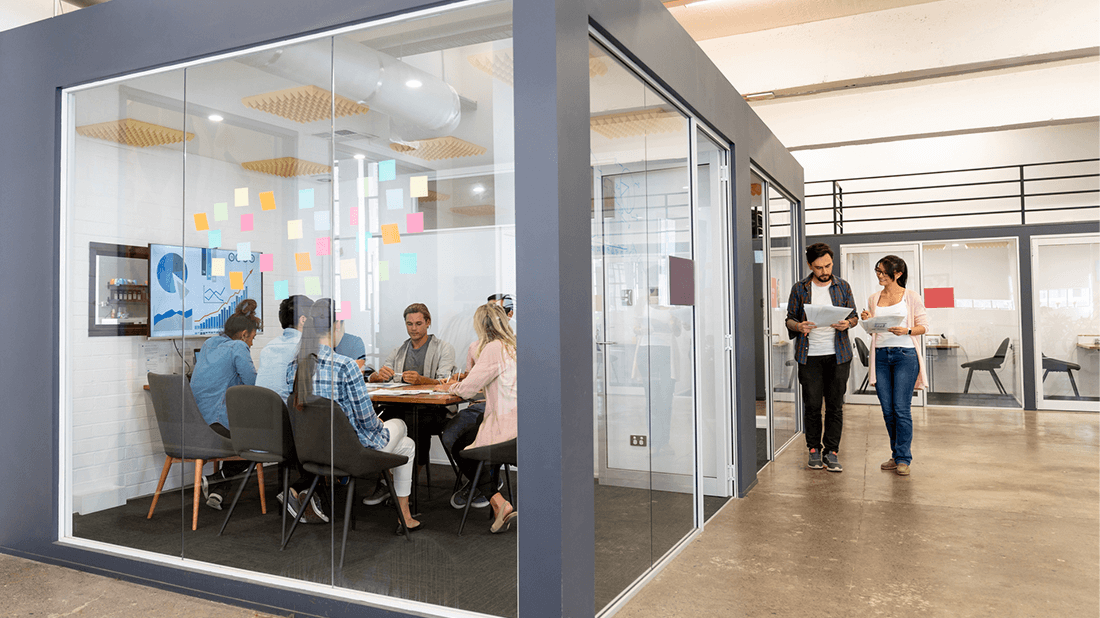 Office workers in a small conference room with two people standing outside